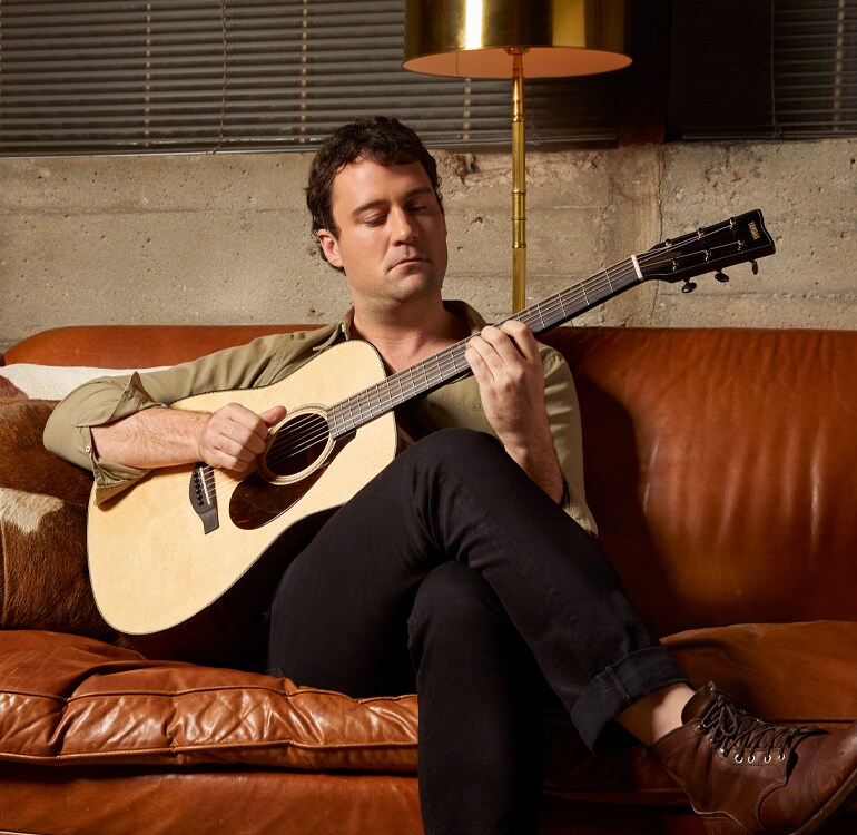 Guitarist Jordan Tice sits on a dark brown leather couch in a room lit with relaxing evening light playing an FG9 acoustic guitar.